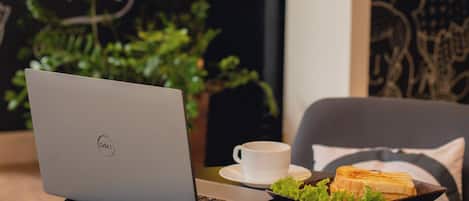 A laptop on a table with a cup and a plate of food, in a cafe with a wall featuring text and a plant.