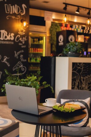 A laptop on a table with a cup and a plate of food, in a cafe with a wall featuring text and a plant. - ibis budget BH Afonso Pena Savassi (Belo Horizonte)