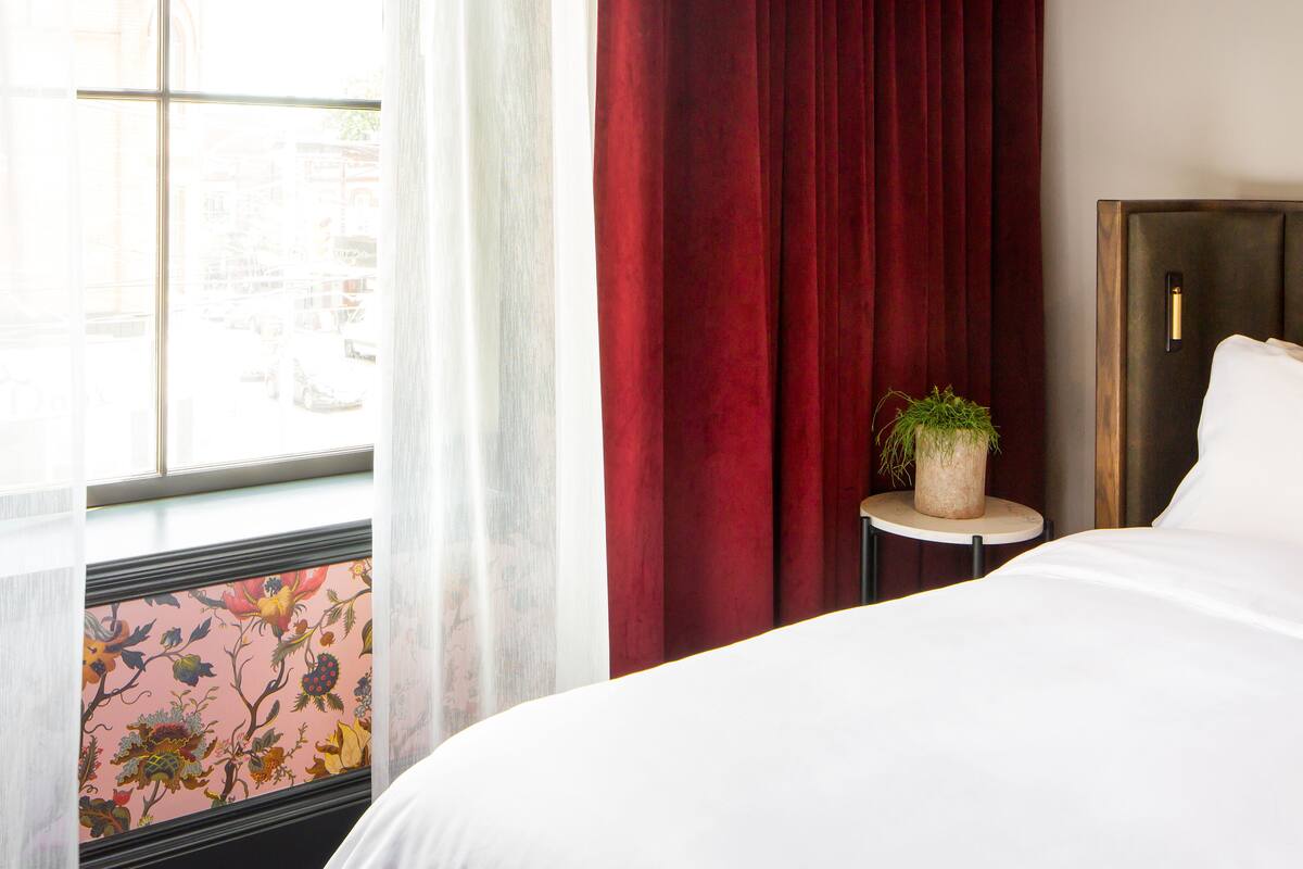 Habitación de hotel con una cama grande, una mesita redonda con una planta, una ventana con cortinas translúcidas y una cortina roja.