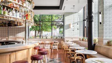 A modern bar area with a variety of liquor bottles, wooden bar counters, and stools. There are multiple seating areas with tables and chairs, and large windows providing a view of the outdoors.