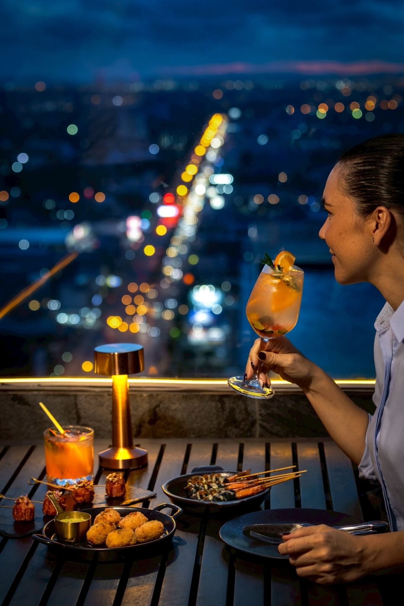 A person holding a cocktail glass with a garnish, seated at a table with a view of city lights.