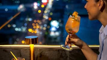 A person holding a cocktail glass with a garnish, seated at a table with a view of city lights.