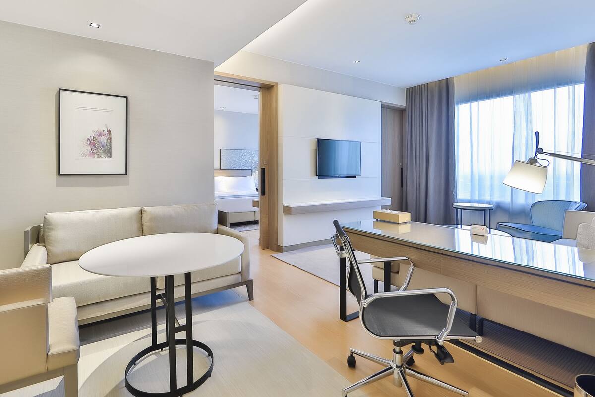 A modern hotel room with a sofa, a round table, a desk with a chair, a flat-screen TV, and a large window with curtains.