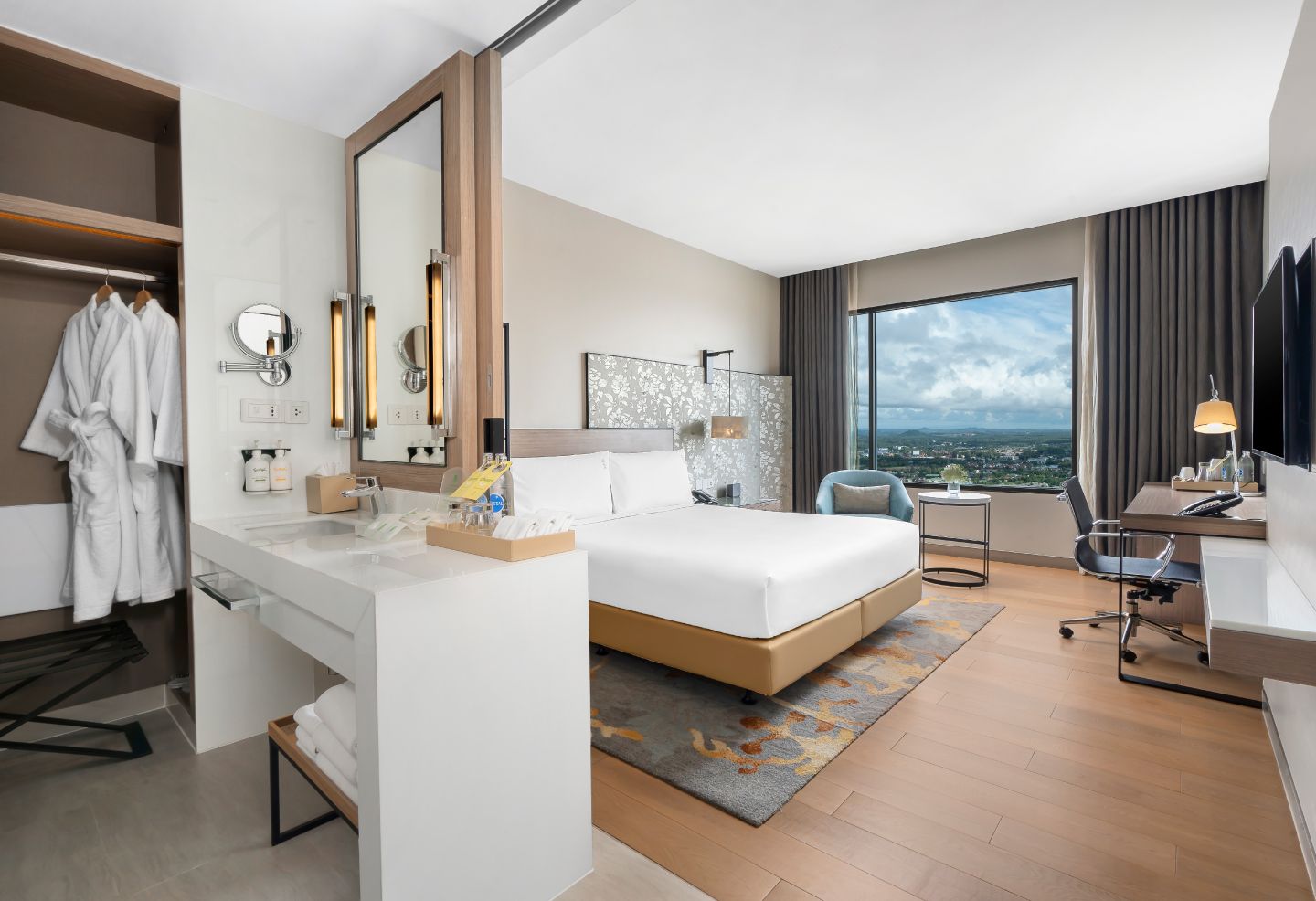 A modern hotel room with a large bed, a desk, a chair, a vanity with a mirror, and a view of the city.