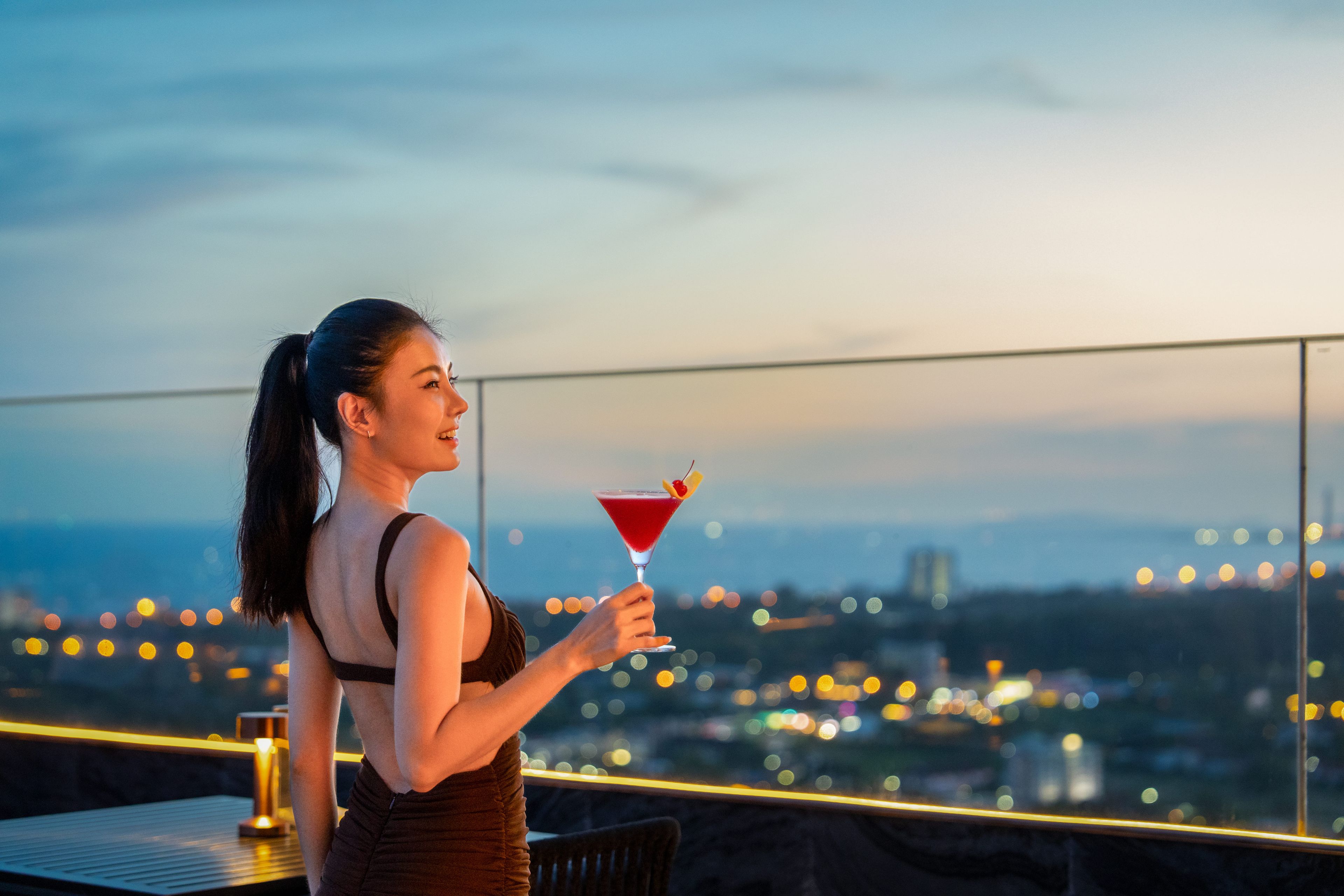 Eine Person hält ein Martini-Glas auf einem Balkon mit Blick auf die Stadt.