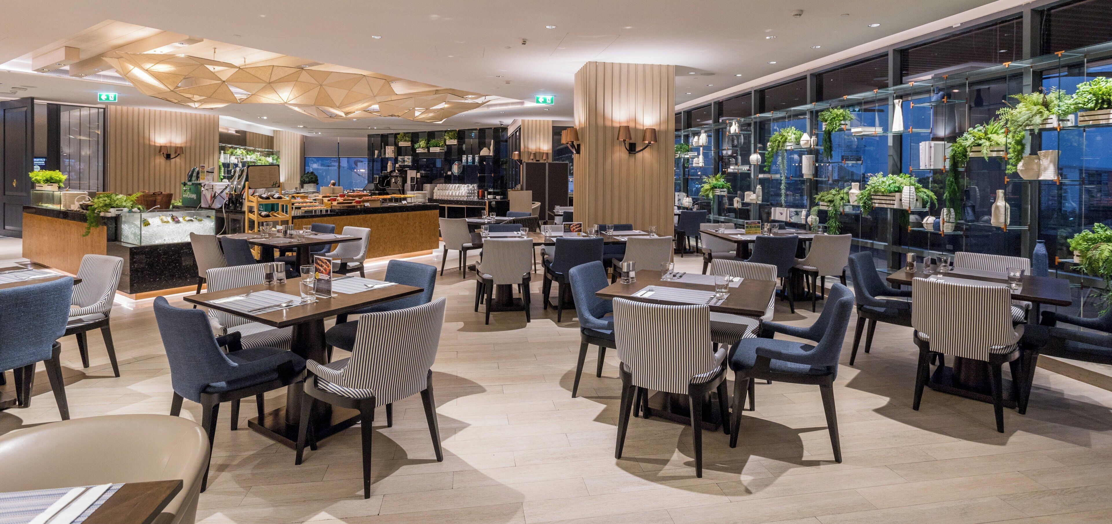 A modern restaurant with a bar area, seating with striped chairs, and a view of the city through large windows.