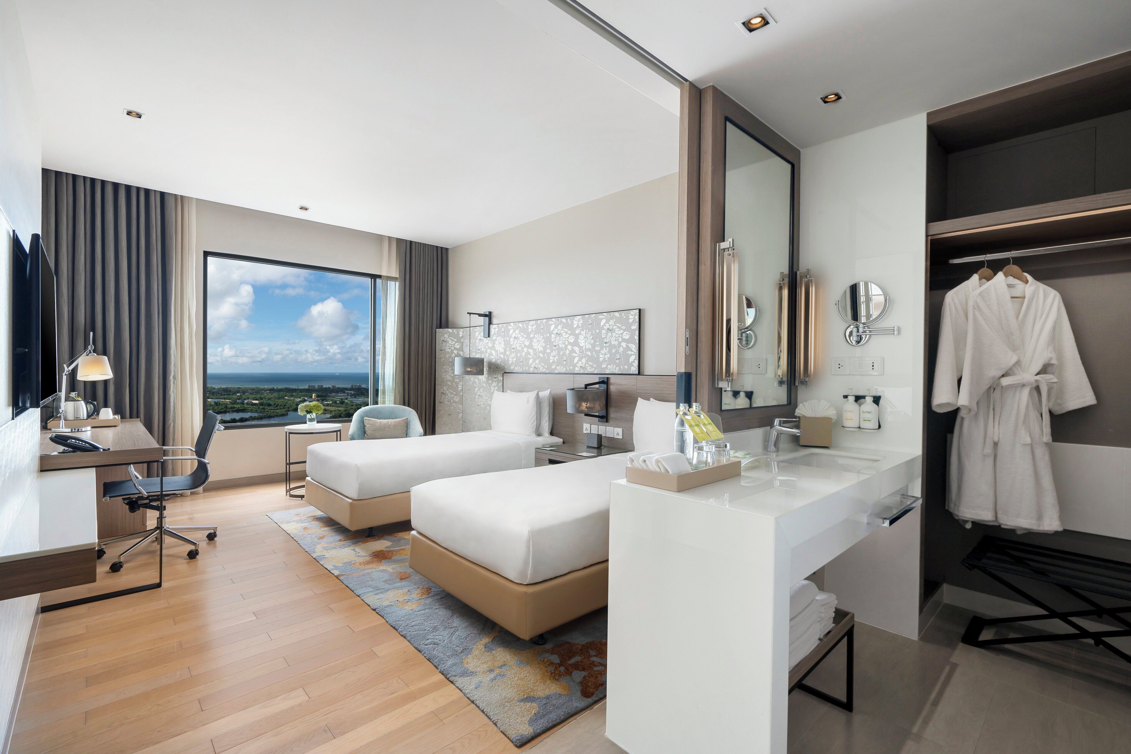 Habitación de hotel con un ventanal grande, un escritorio, dos camas, un baño con espejo y una bata colgada en la pared.