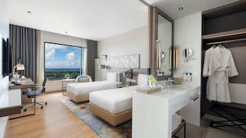 A hotel room with a large window, a desk, two beds, a bathroom with a mirror, and a robe hanging on the wall.
