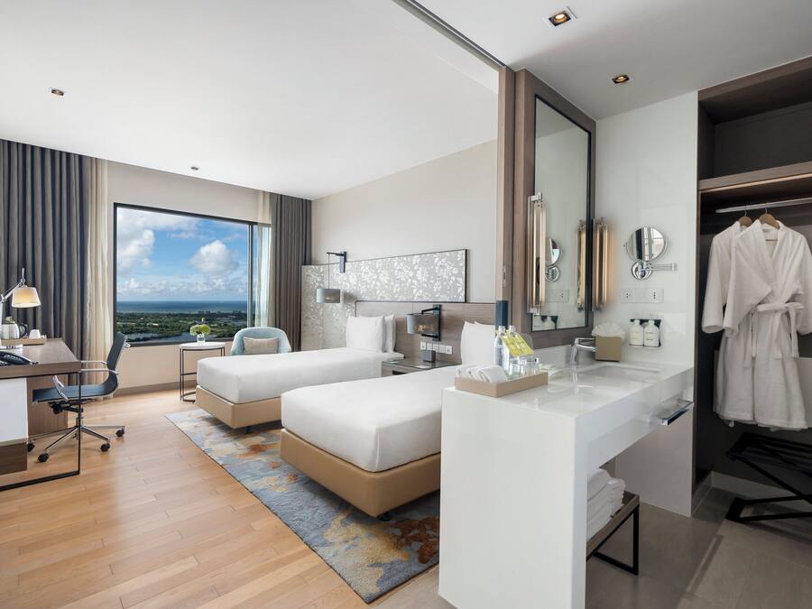 A hotel room with a large window, a desk, two beds, a bathroom with a mirror, and a robe hanging on the wall.