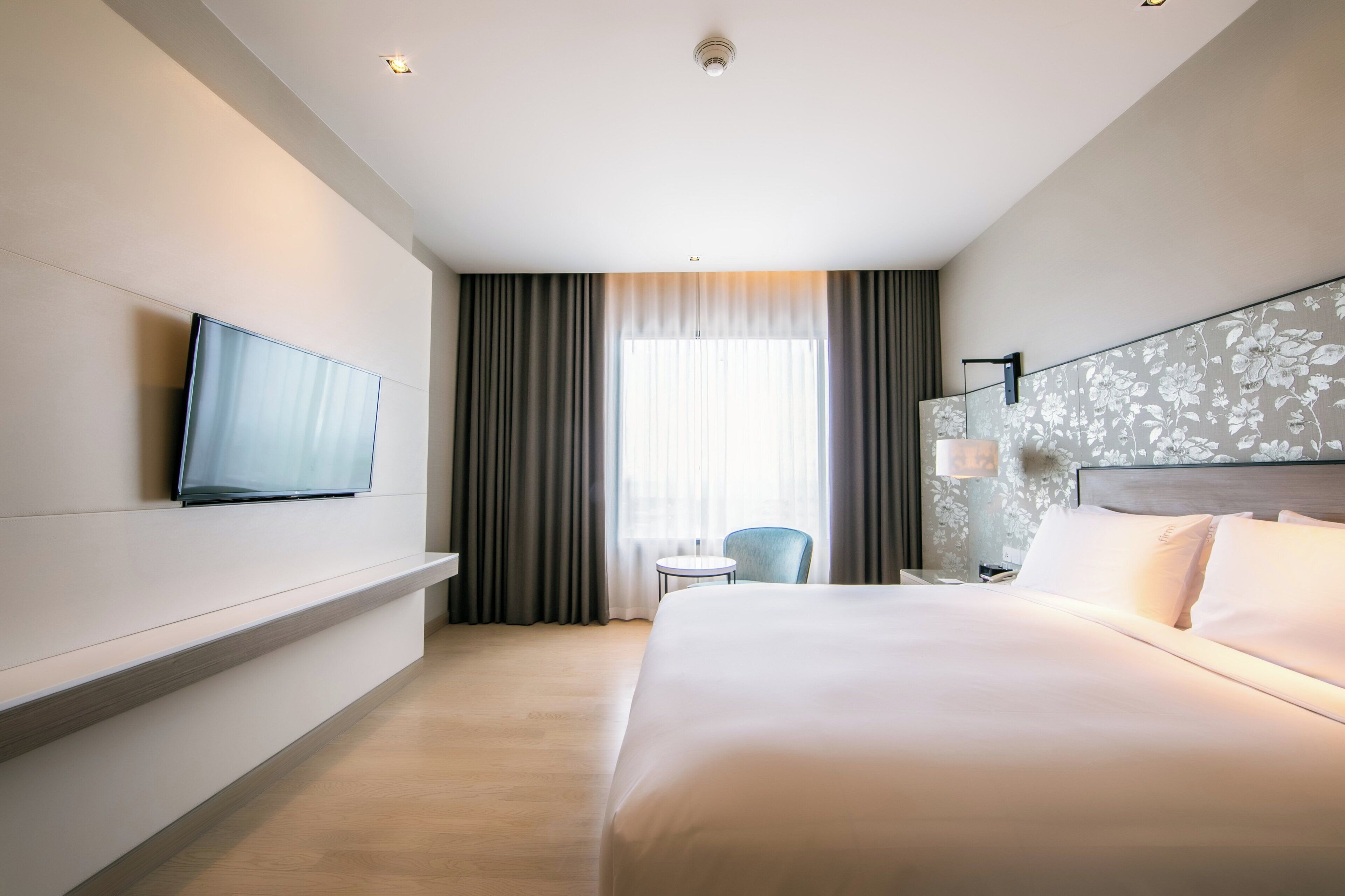 A hotel room with a bed, a television mounted on the wall, a small table, a chair, and a window with curtains.