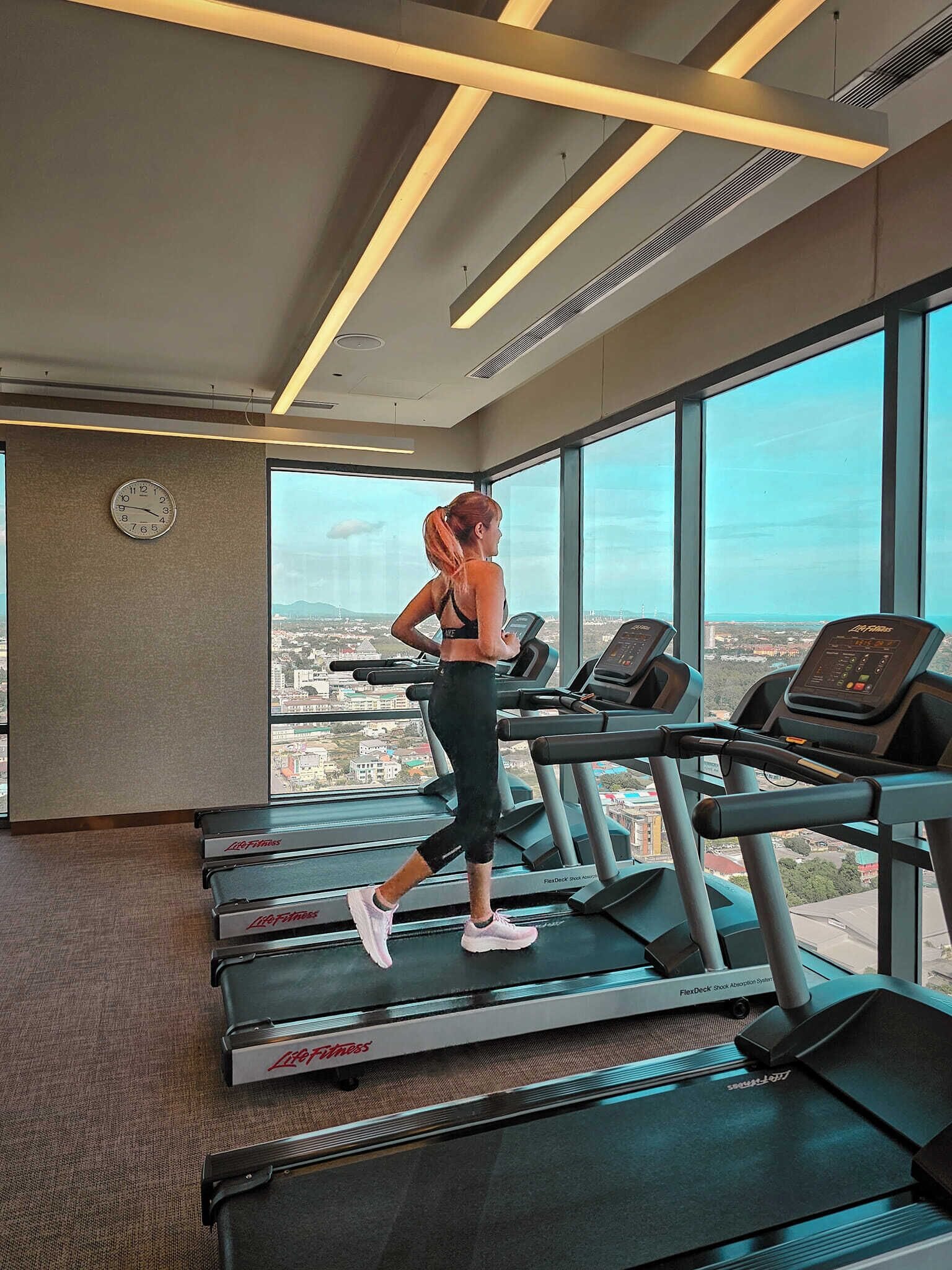 Un gimnasio con cintas de correr, un ventanal con vistas a la ciudad y un reloj de pared.