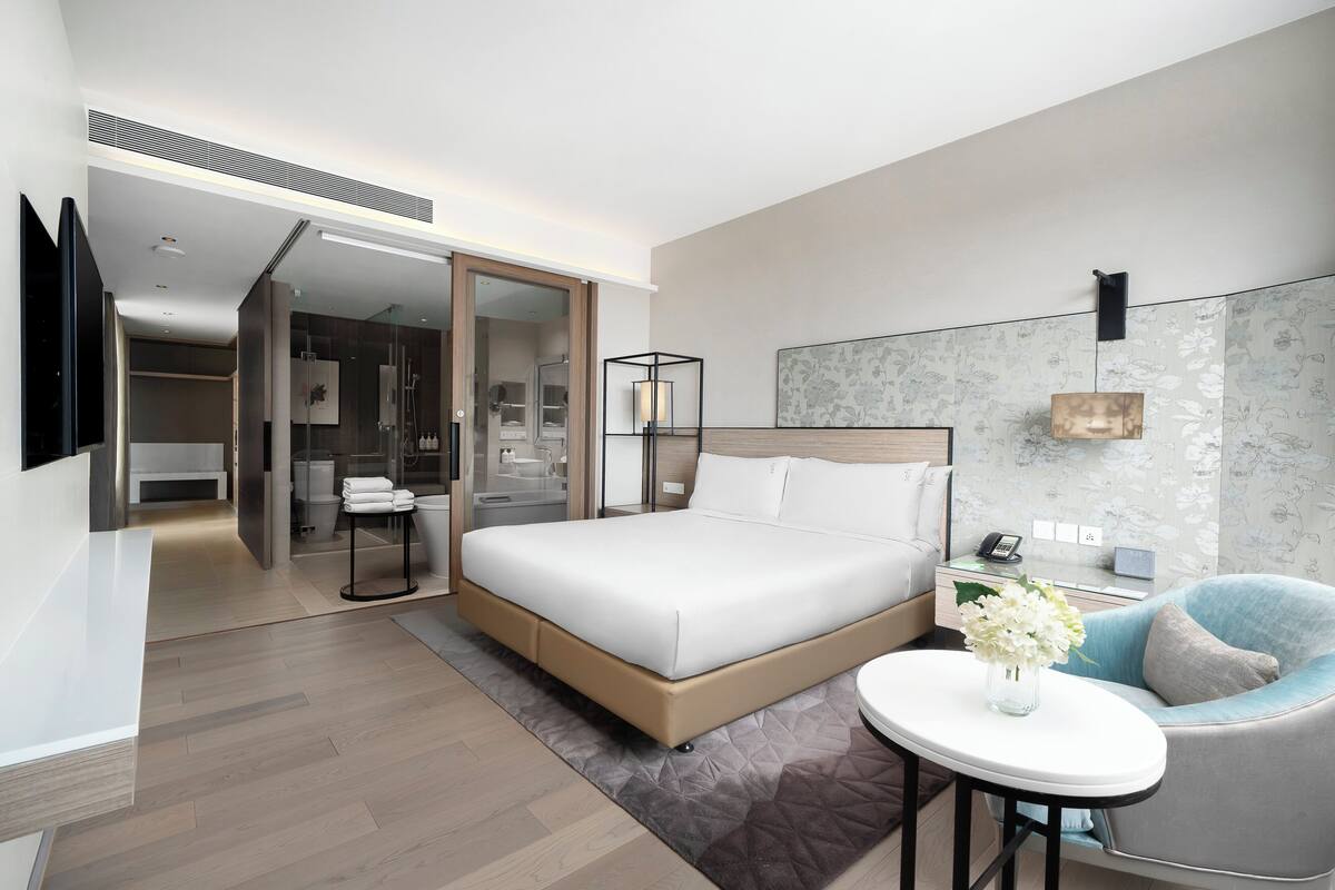 A modern hotel room with a large bed, a small round table with a vase of flowers, a chair, and a desk with a lamp.
