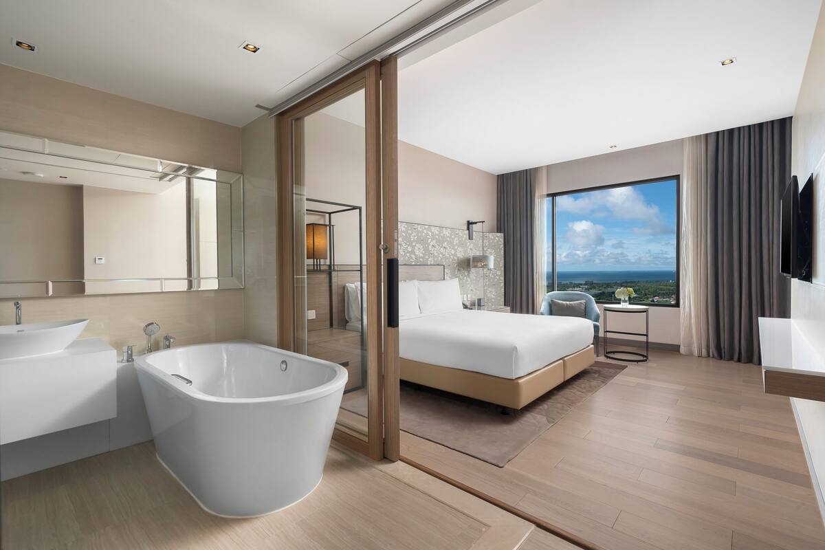 A modern hotel room with a large bathtub, a bed, a desk, and a view of the ocean.