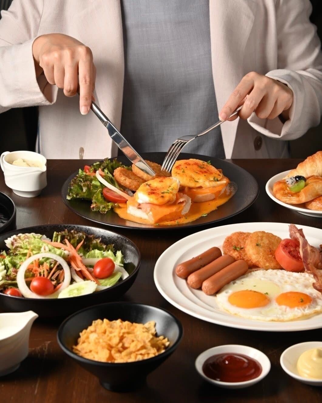 Una persona comiendo una variedad de platos, incluyendo huevos, salchichas y una ensalada.