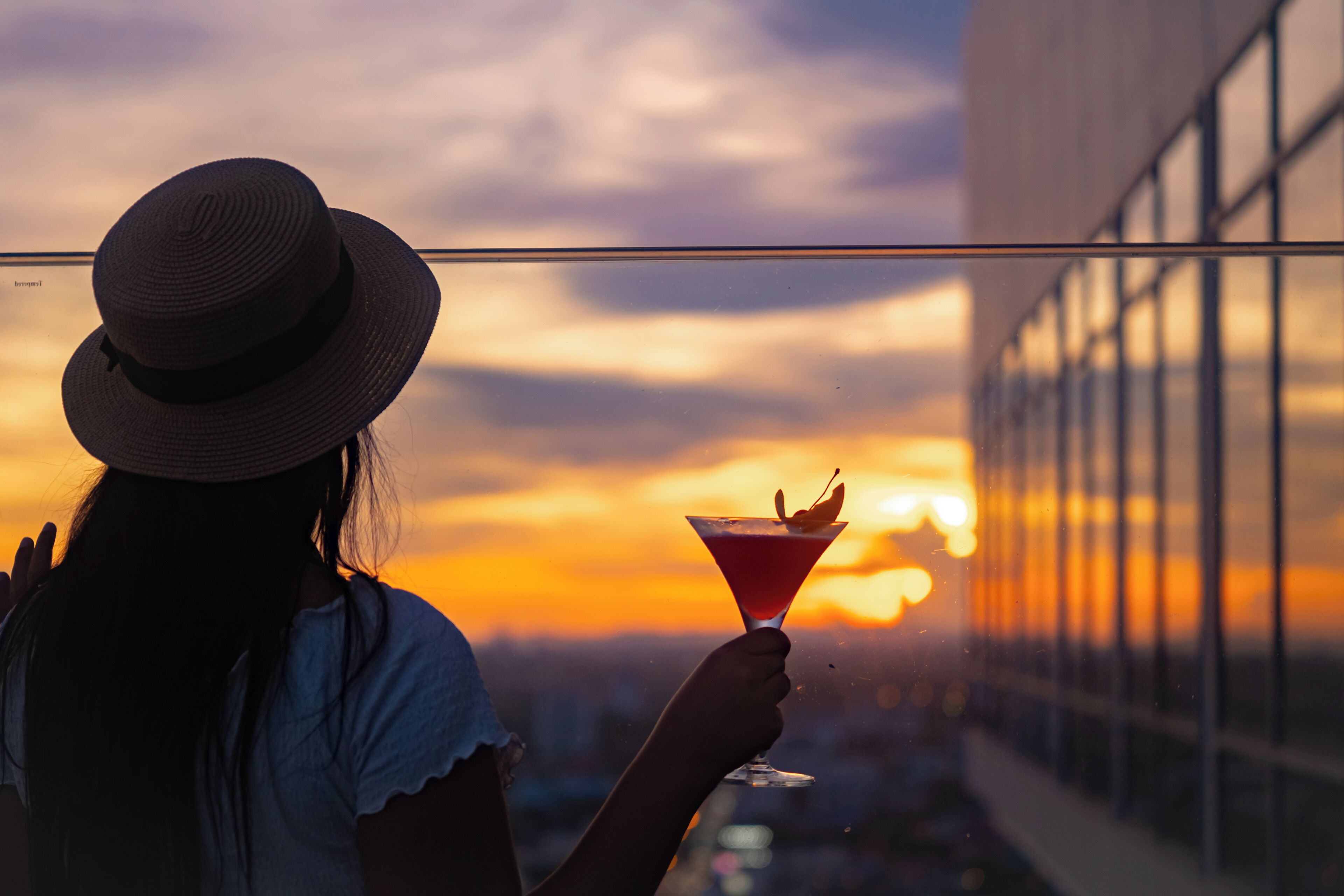 A person holding a cocktail glass with a cherry garnish, against a sunset backdrop.