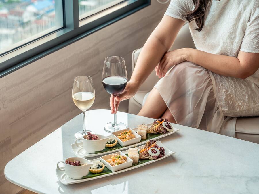 A person seated by a table with a wine glass and a tray of appetizers.