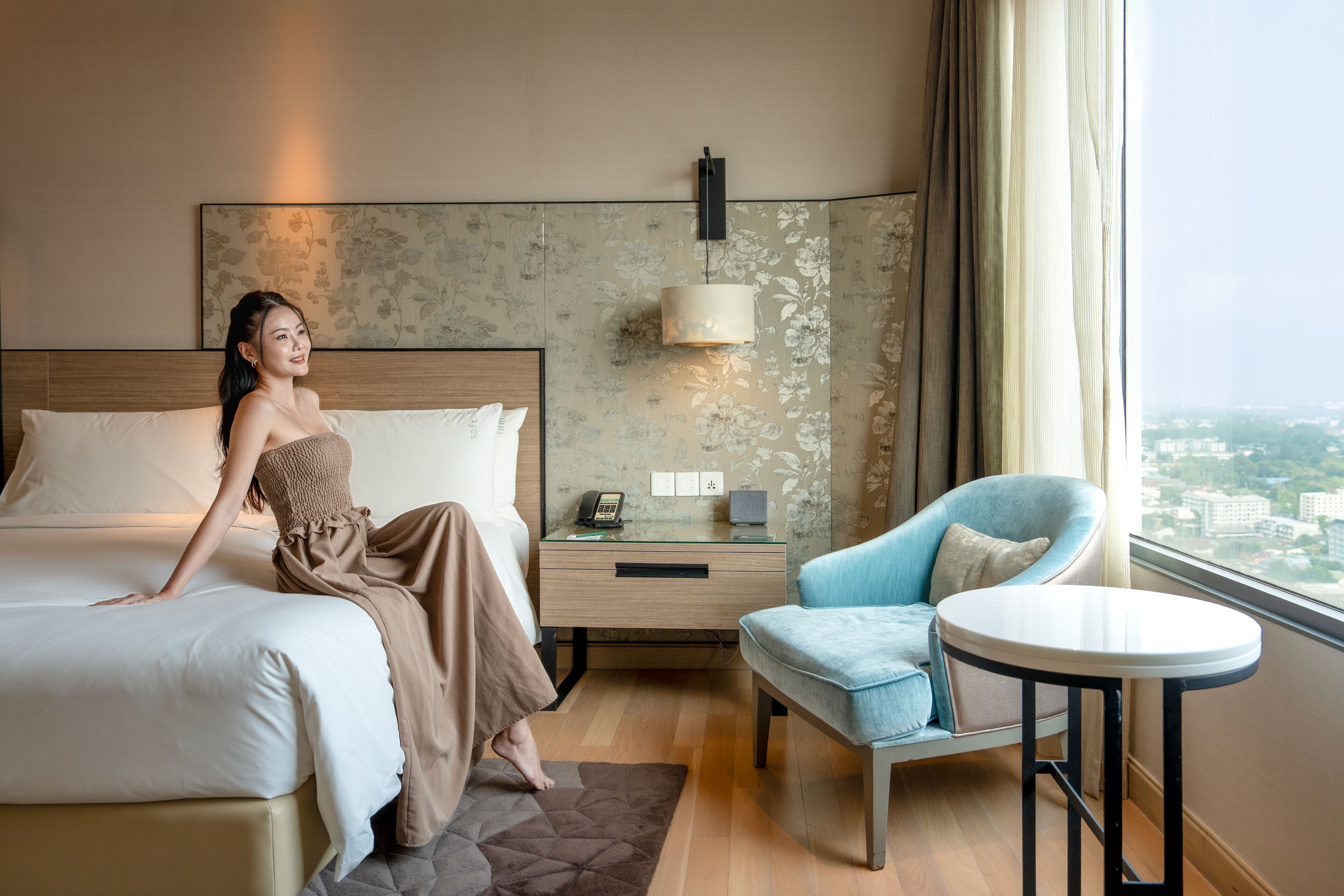 Quarto de hotel com cama, escrivaninha com luminária, cadeira e vista para a cidade.