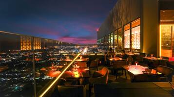 Un espacio de comedor en la azotea con mesas y sillas, con vista a un paisaje urbano al atardecer.