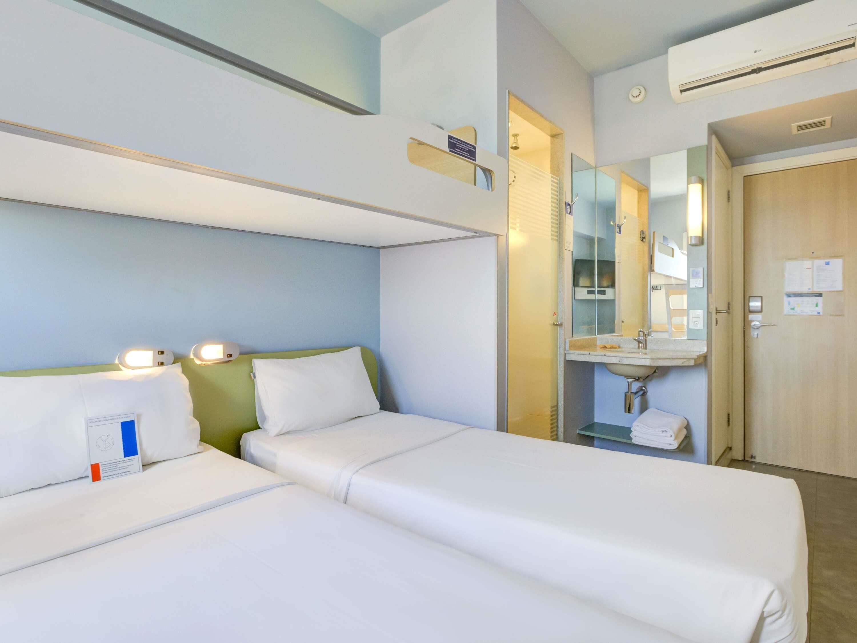 Une chambre d’hôtel avec deux lits simples, une salle de bain avec un lavabo et un miroir, et une porte avec une serrure.