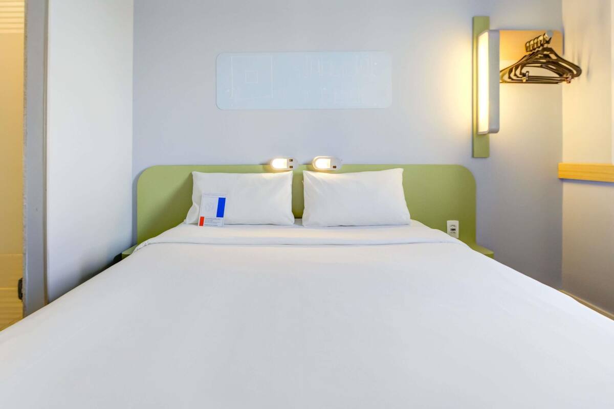 Une chambre d’hôtel équipée d’un lit, d’oreillers, d’une table de chevet, d’une lampe fixée au mur et d’un porte-manteau.