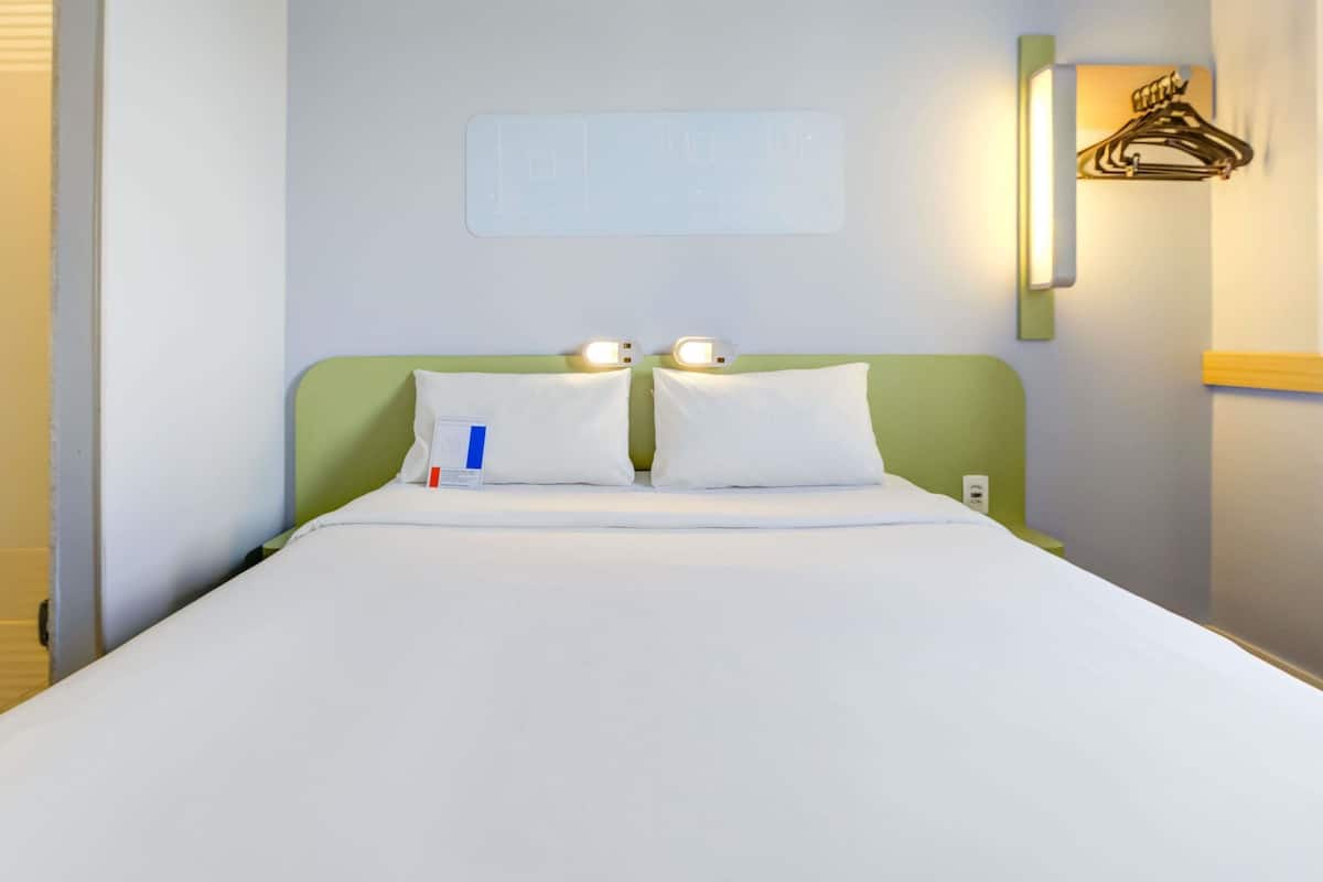 A hotel room with a bed, pillows, a nightstand, a wall-mounted light, and a coat hook.