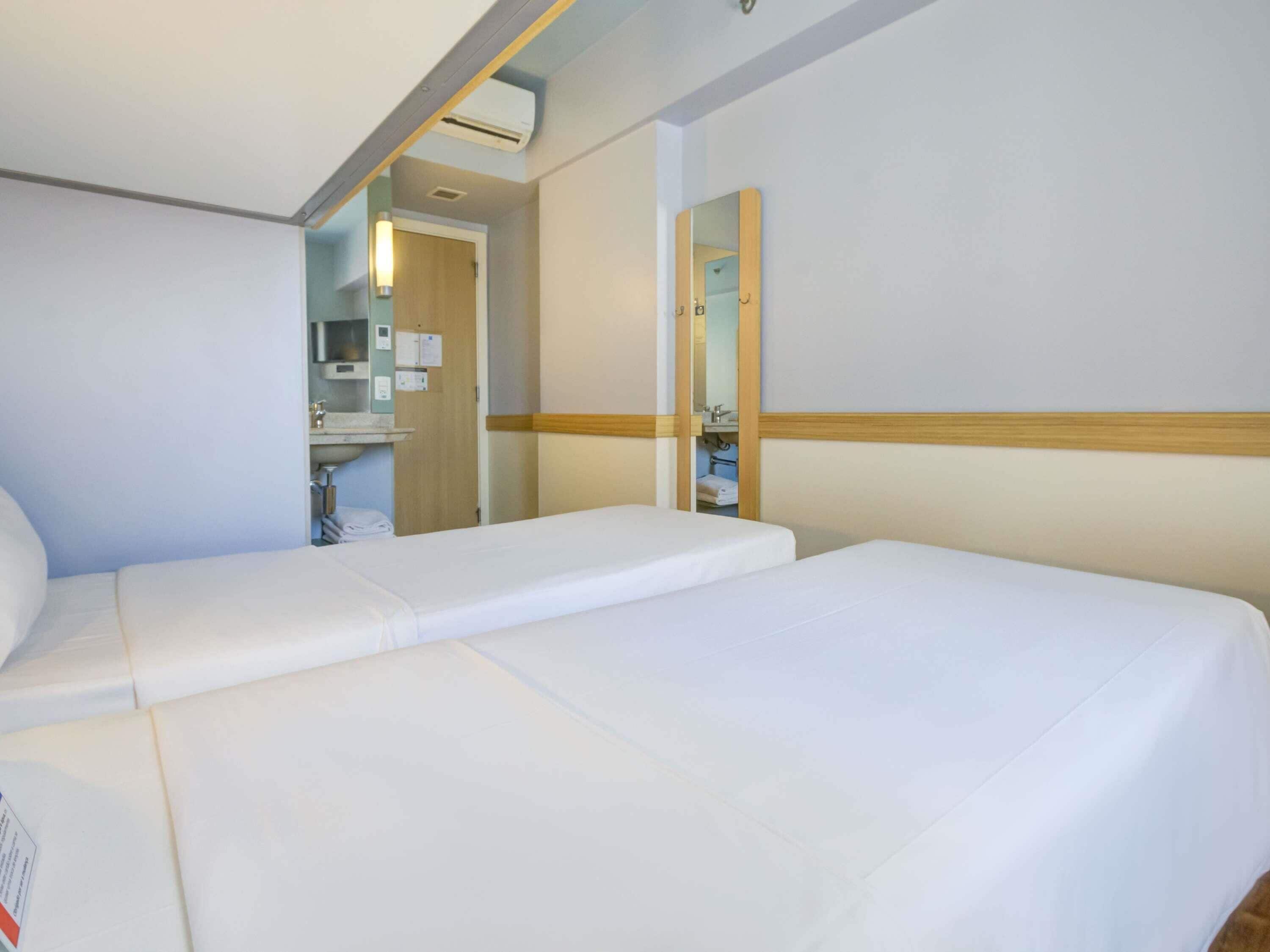 A hotel room with two single beds, a large mirror, and a bathroom visible through an open door.