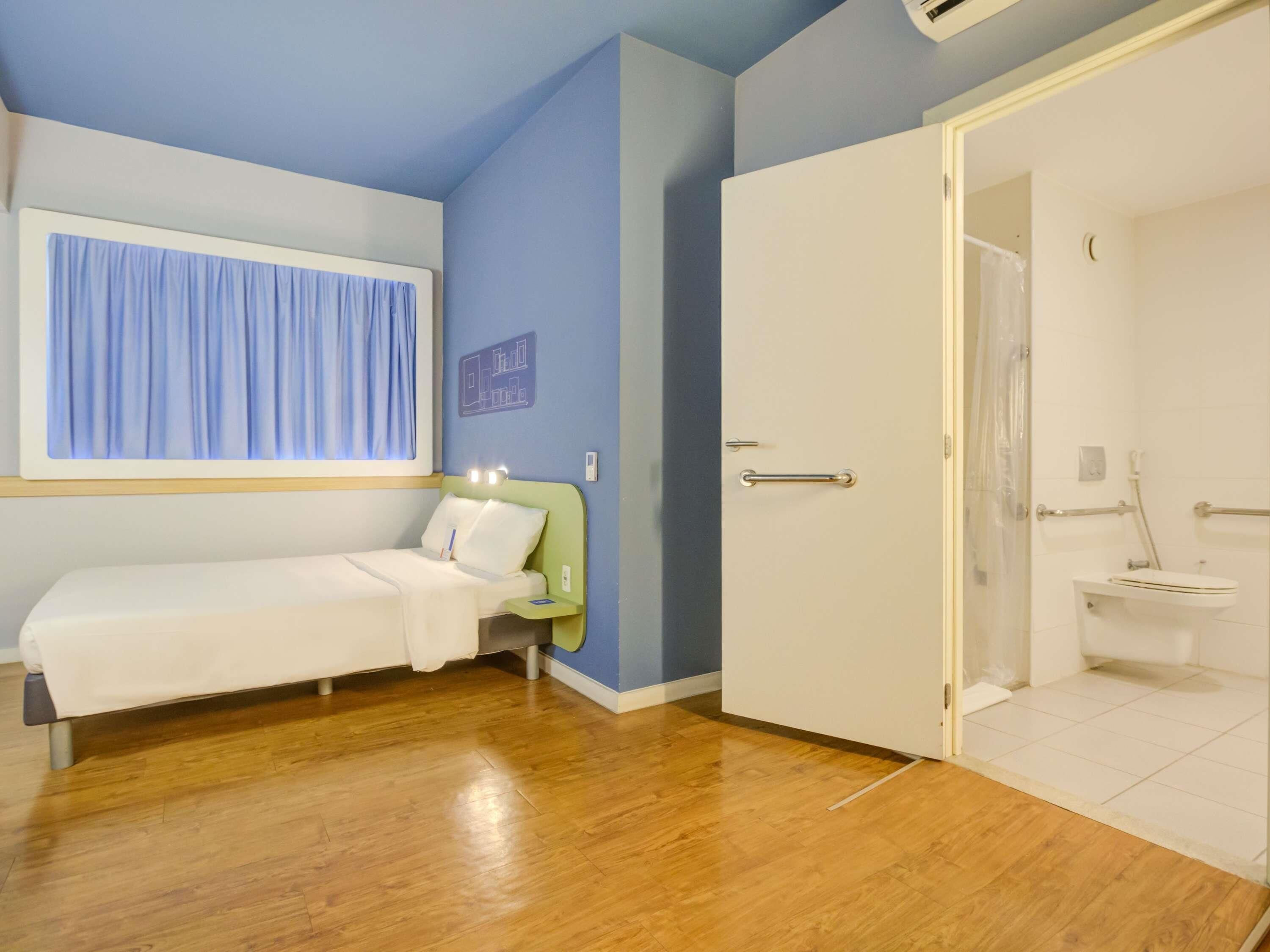 Une pièce comprenant un lit, une fenêtre avec des rideaux, une porte donnant sur une salle de bain et un climatiseur fixé au mur.