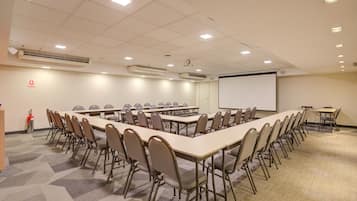 Une salle de conférence équipée de longues tables et de chaises, d’un écran de projection et d’un extincteur.