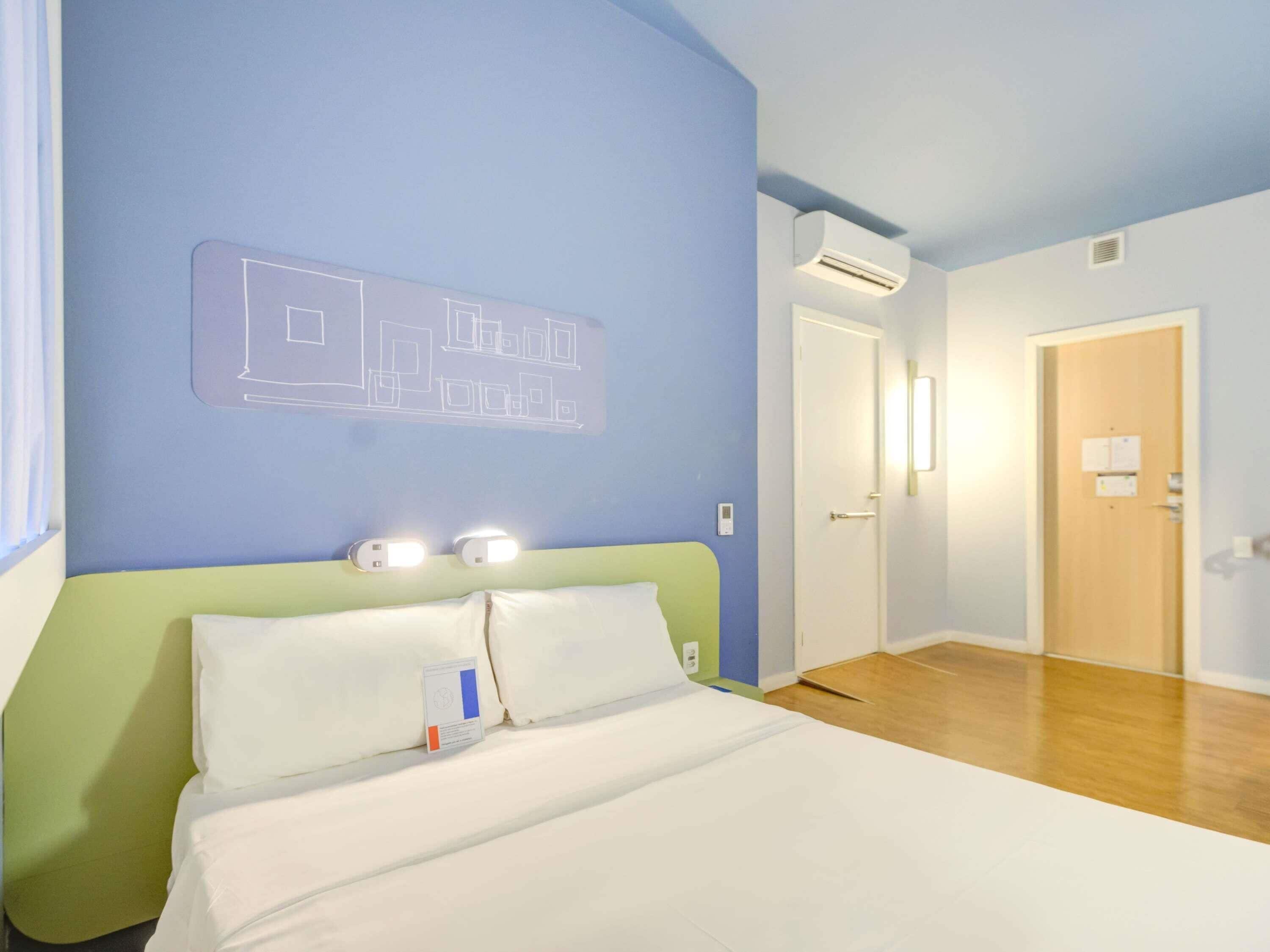 A hotel room with a bed, a headboard, a wall-mounted air conditioner, and a door.