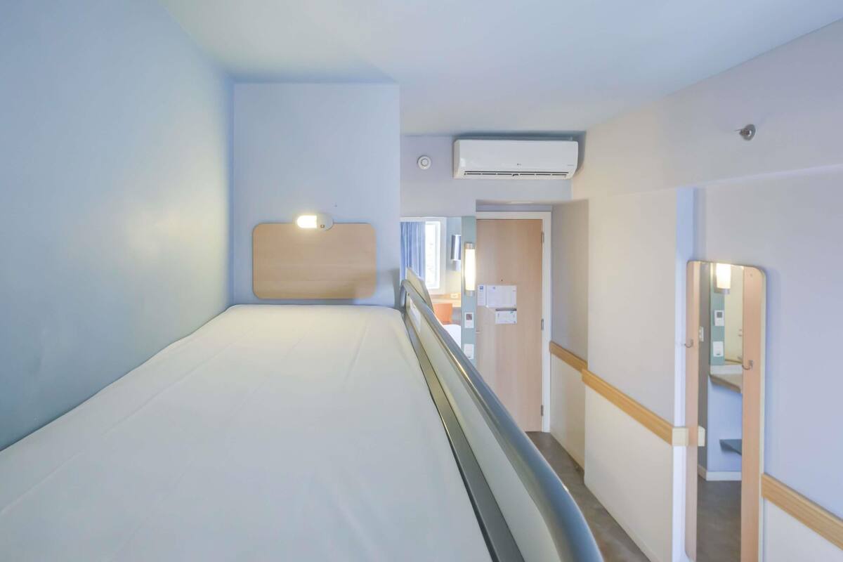 Ett hotellrum med en säng, en sänggavel, en luftkonditioneringsanläggning, en spegel och en dörr som leder till ett badrum.