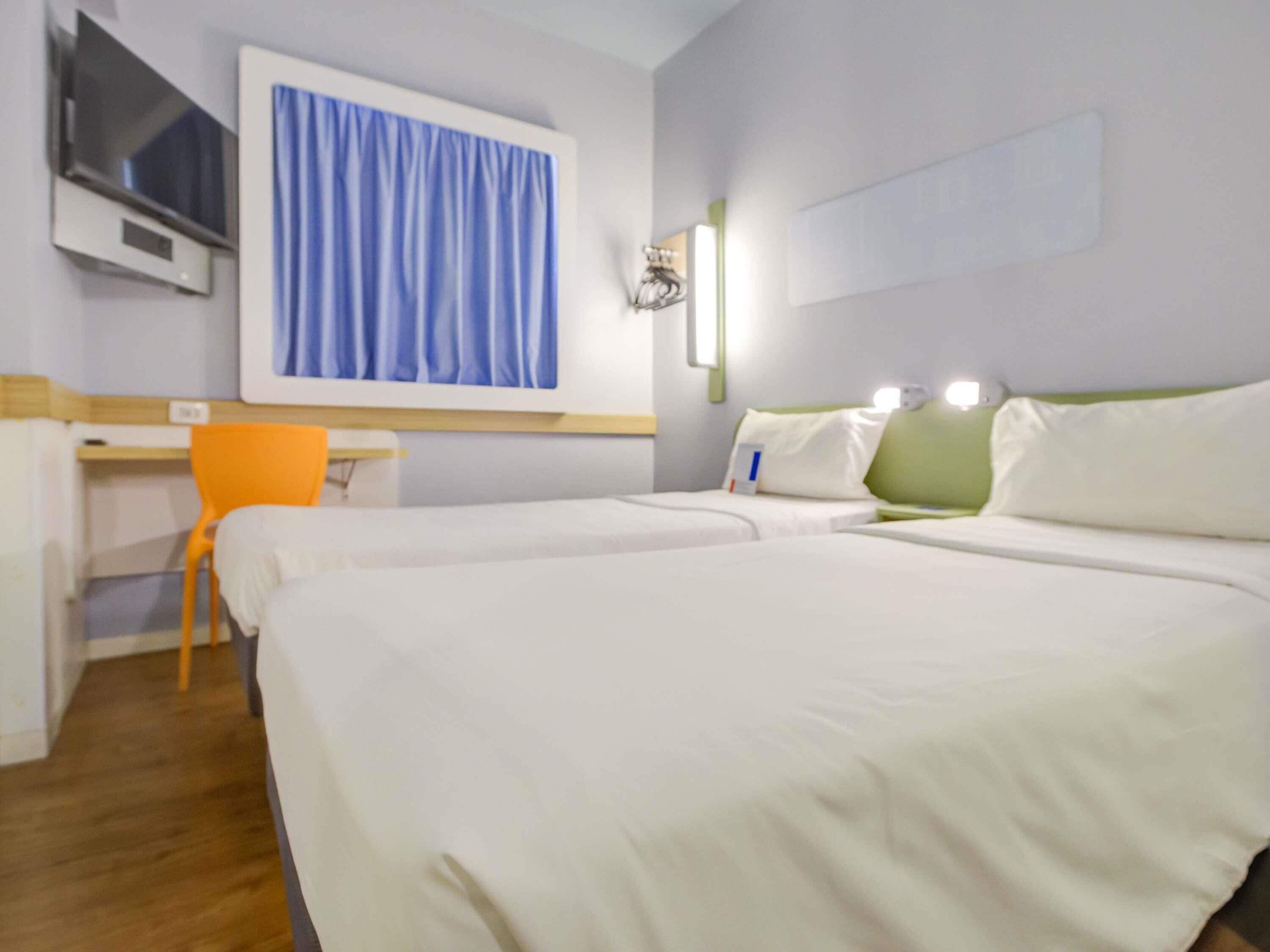 A hotel room with two beds, a desk, a chair, a TV, and a window with curtains.