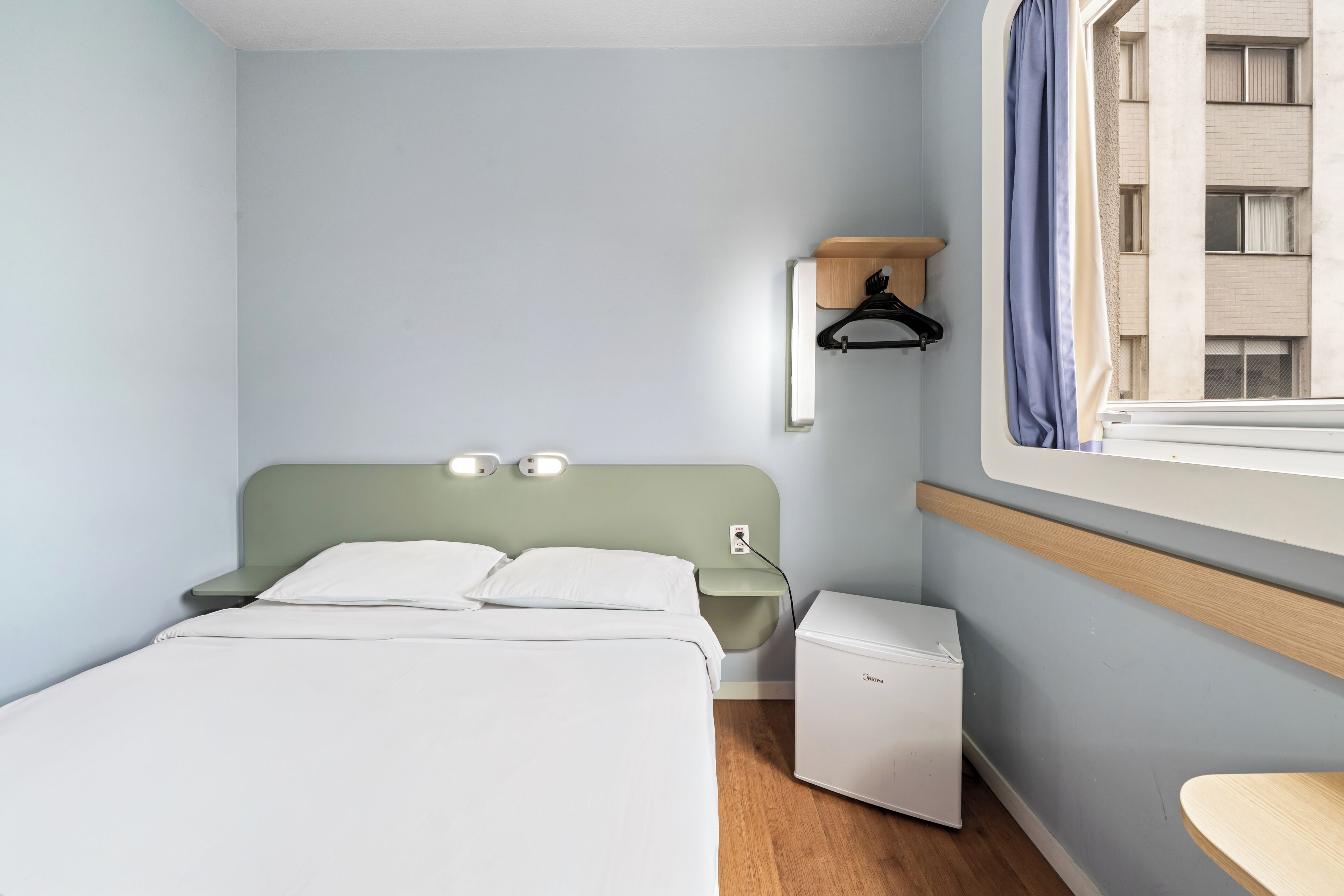 A hotel room with a bed, bedside table, nightstand, and a small refrigerator.