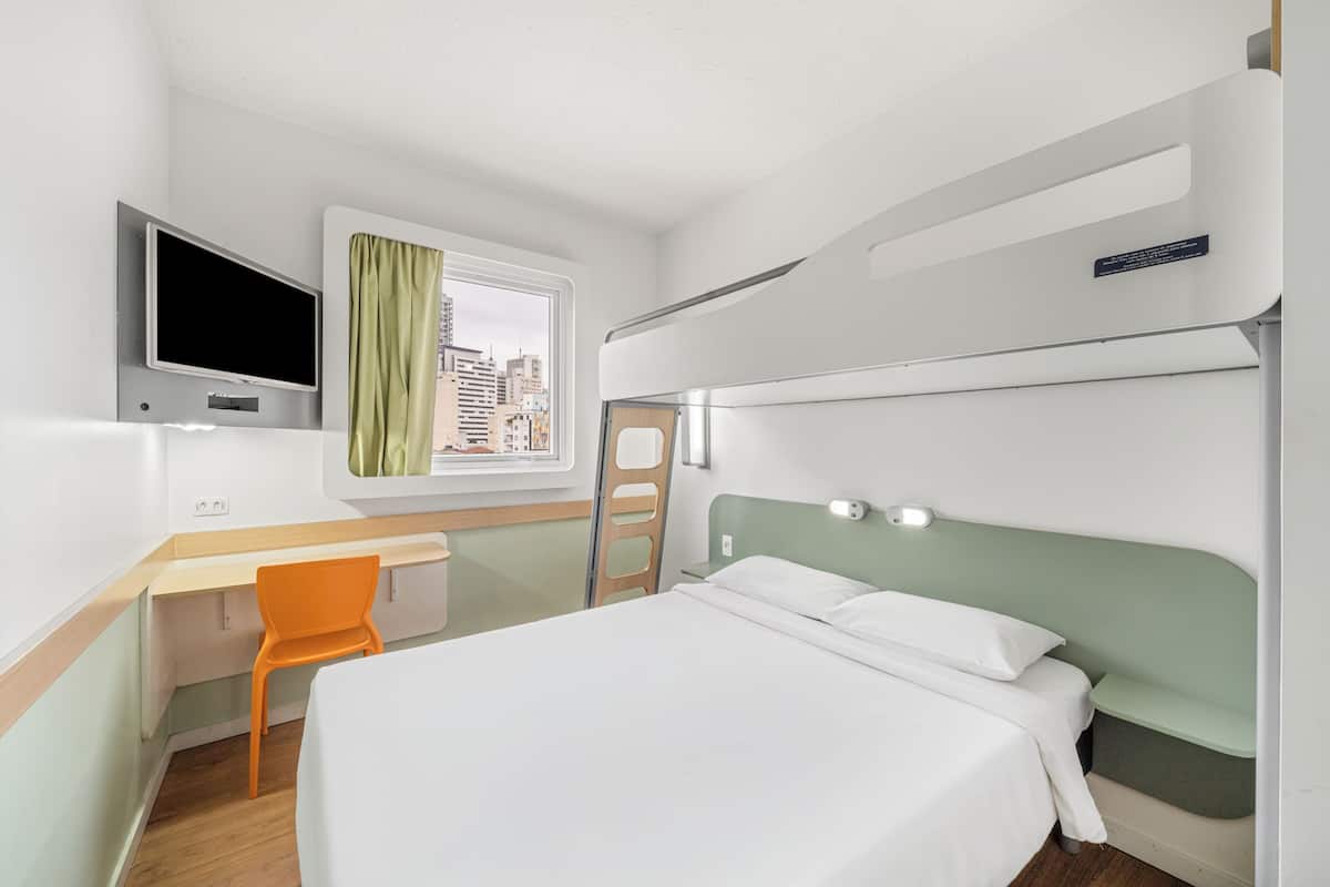 A hotel room with a bed, desk, chair, TV, window with curtains, and a city view.