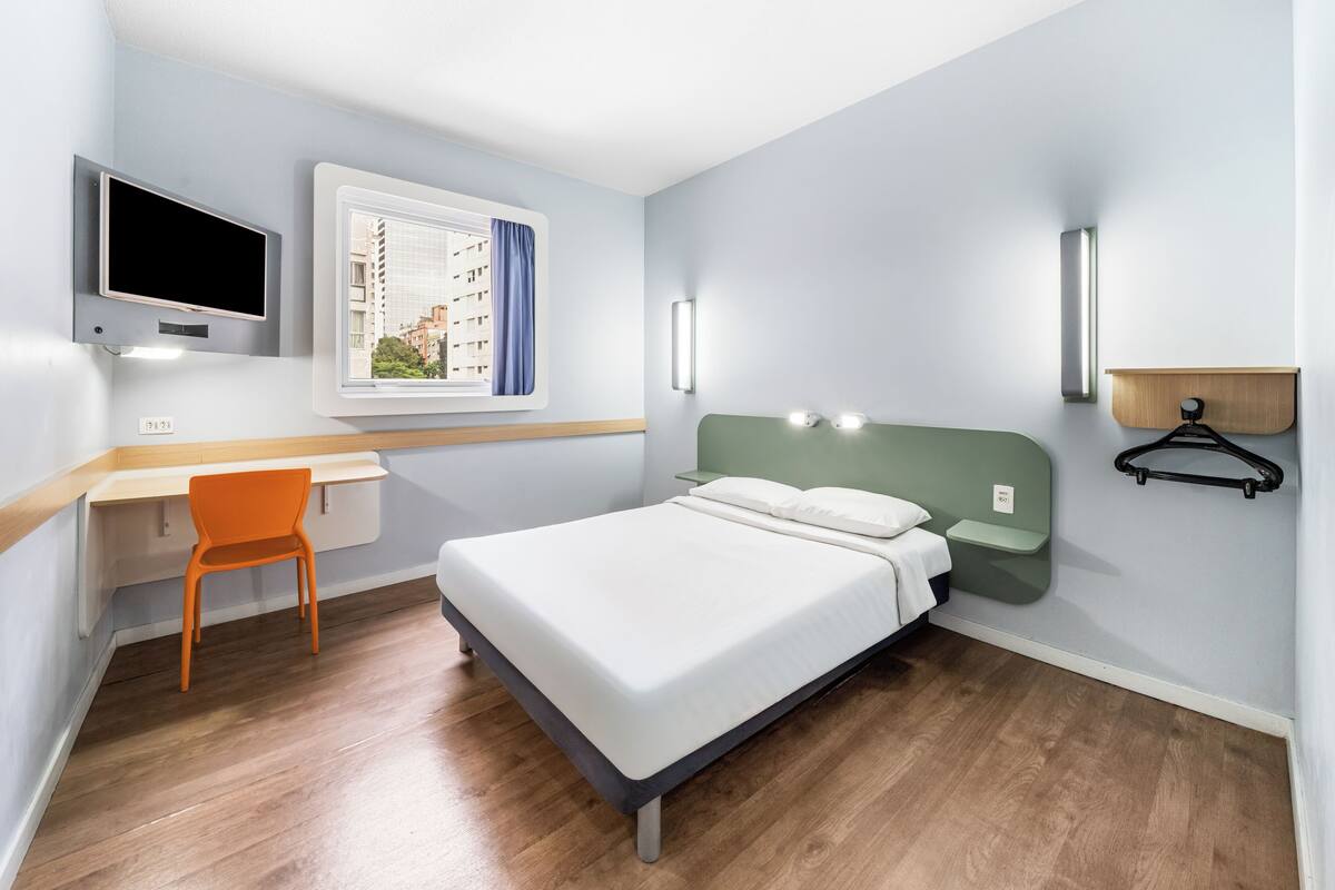 A bedroom with a bed, a desk, a chair, a TV, and a window with curtains.