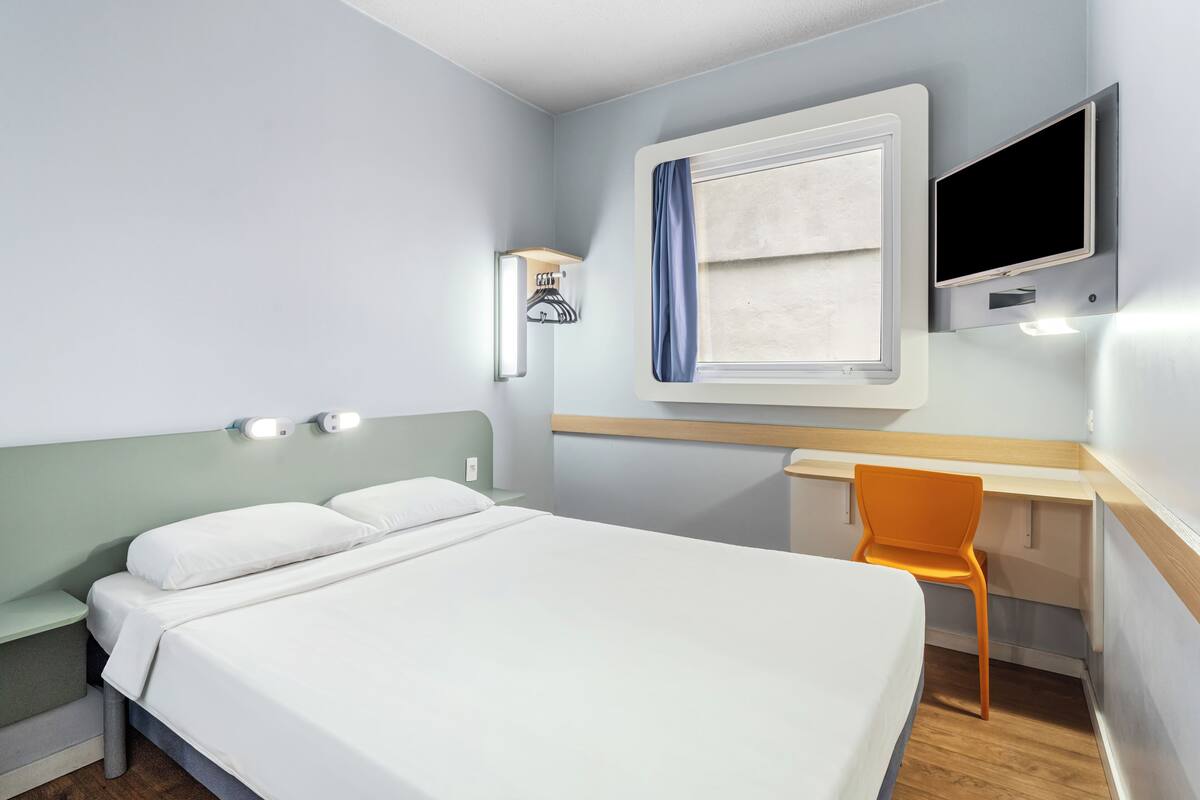 A hotel room with a bed, a desk, a chair, a TV, and a window with a curtain.
