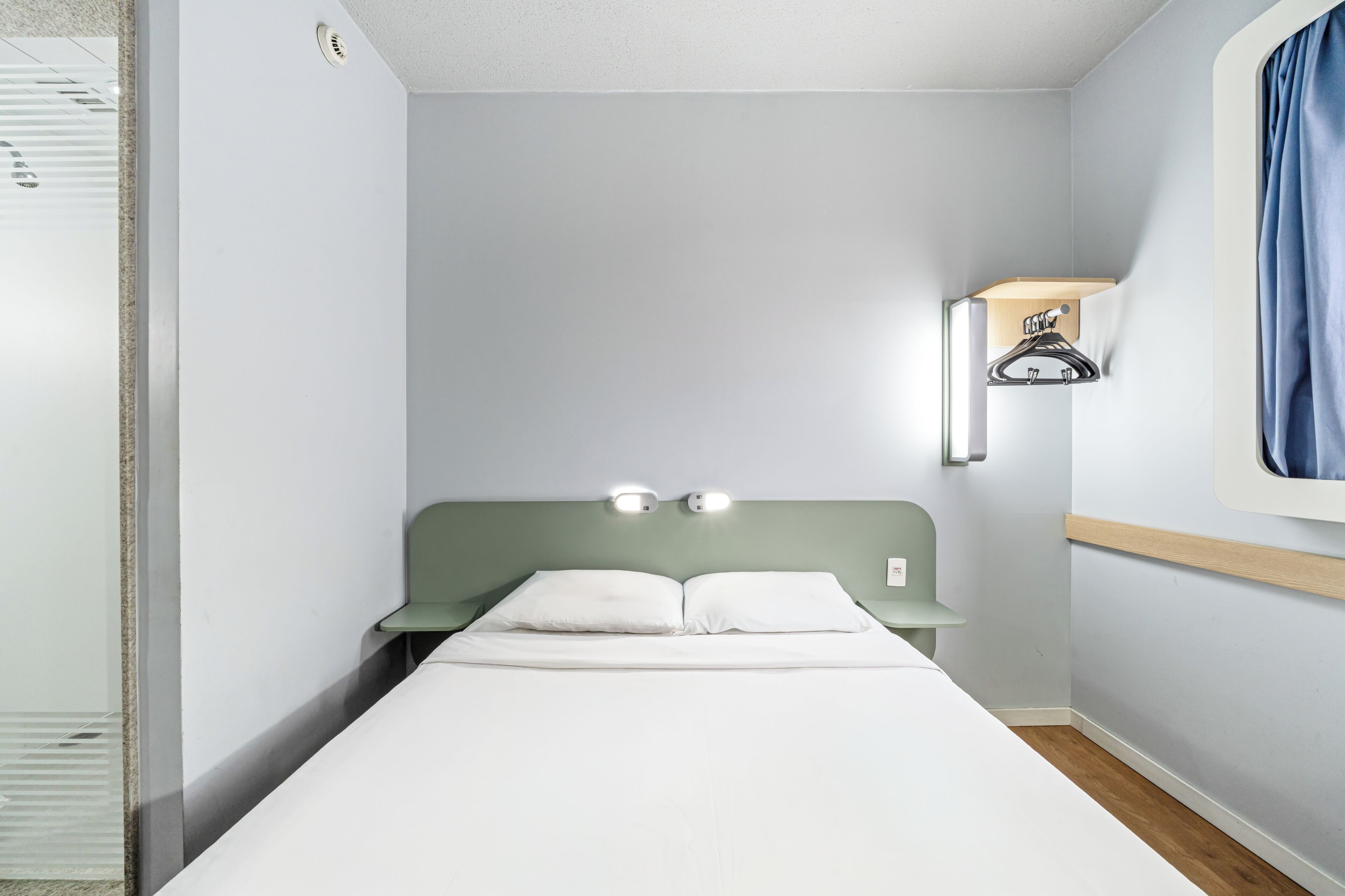 A single bed with a headboard, a nightstand, a wall-mounted shelf with a towel, and a window with curtains.