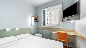 A hotel room with a bed, a desk, a chair, a TV, a window with curtains, and a wall-mounted shelf.