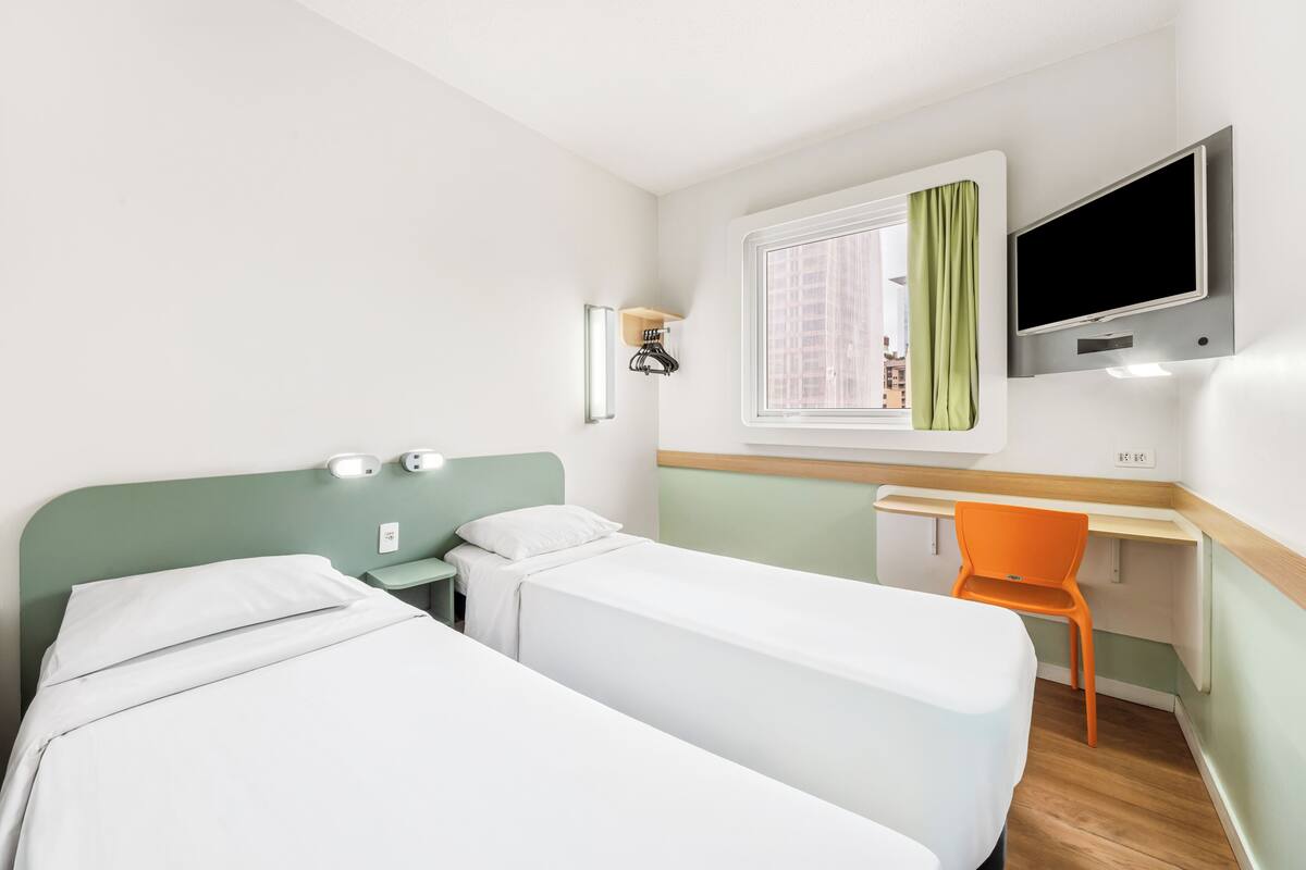 A hotel room with two beds, a desk, a chair, a TV, and a window with curtains.