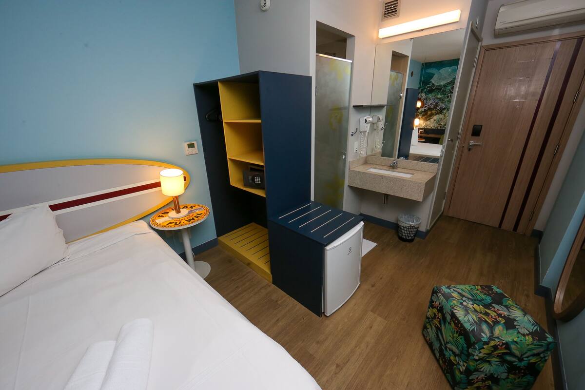 A hotel room with a bed, a small desk with a lamp, a wardrobe, a mirror, a sink, and a door.