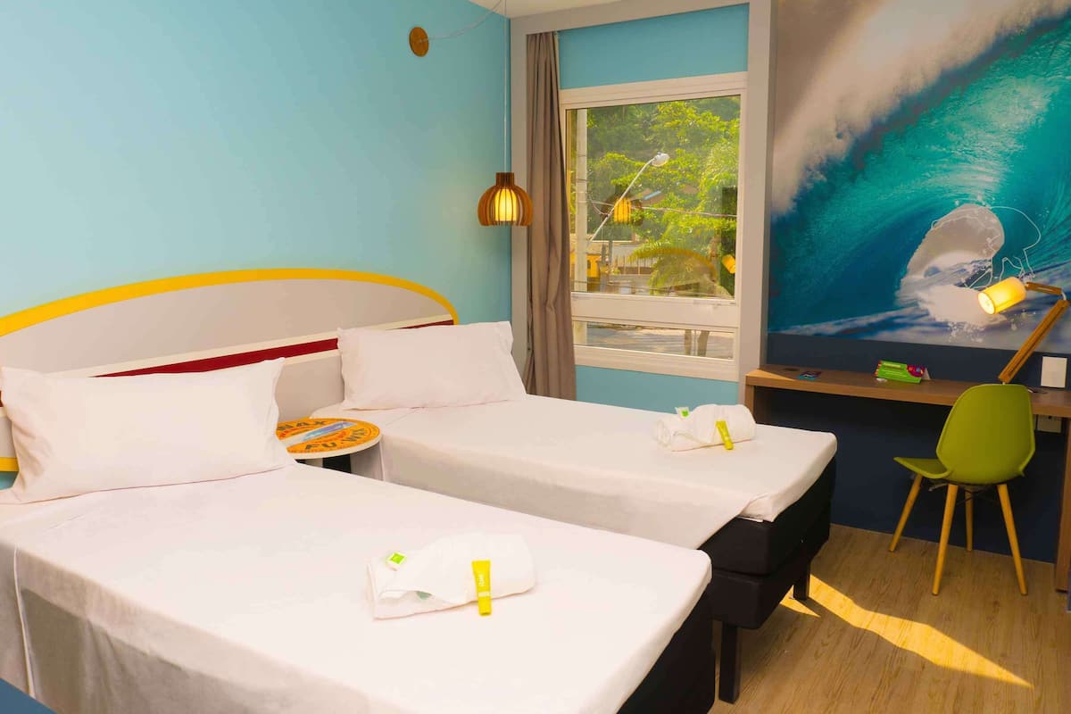 A hotel room with two beds, a desk with a chair, a window with a view of trees, and a wall mural of a wave.