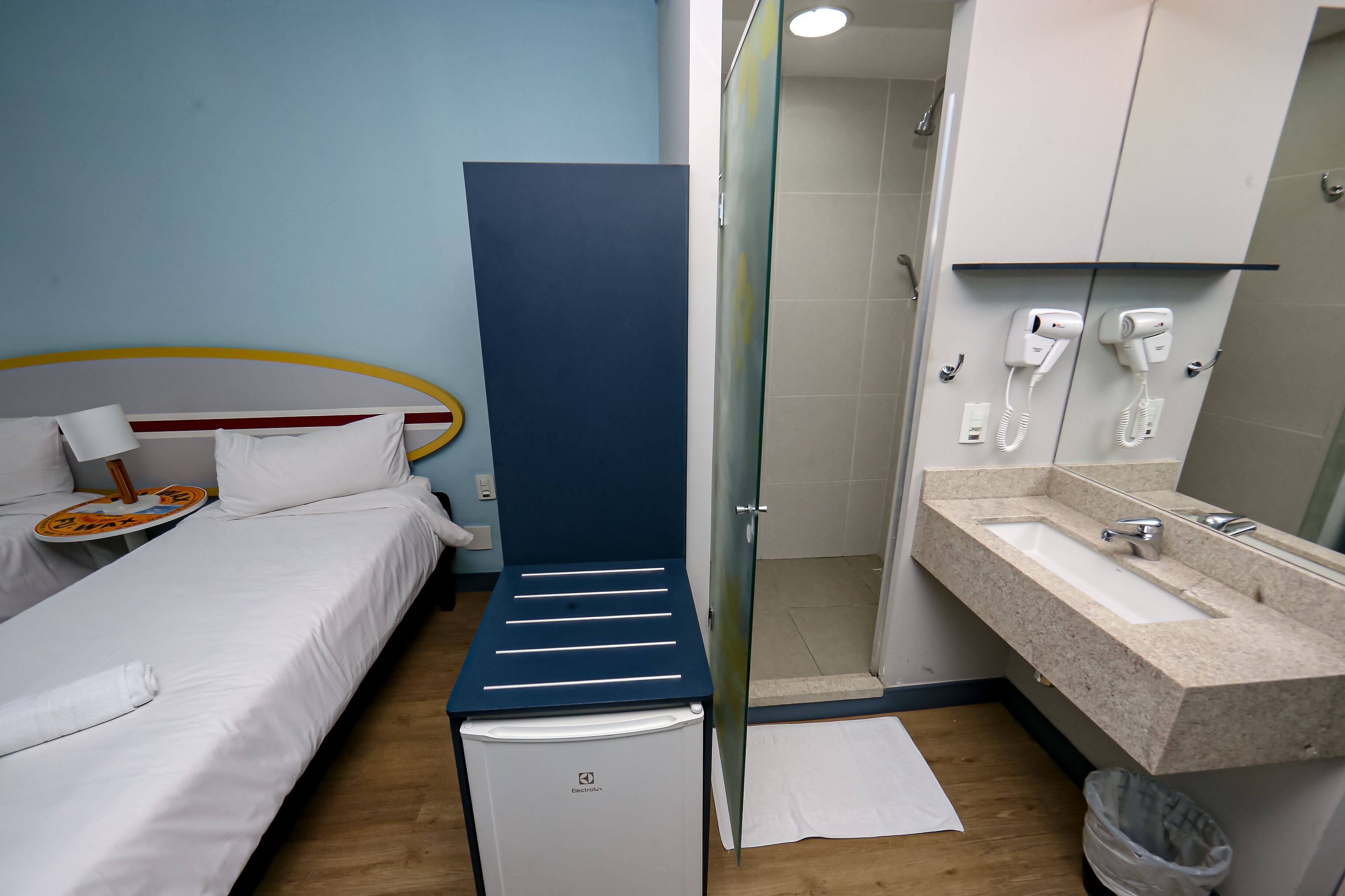 Habitación de hotel con cama, refrigerador pequeño, zona de ducha y lavabo con espejo.