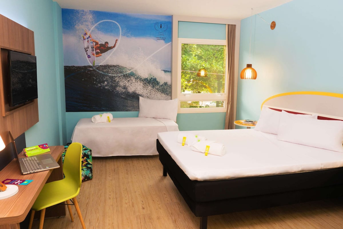 A hotel room with two beds, a desk with a laptop, a chair, a TV, and a mural of a surfer on a wave.