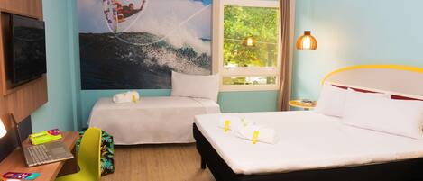 A hotel room with two beds, a desk with a laptop, a chair, a TV, and a mural of a surfer on a wave.
