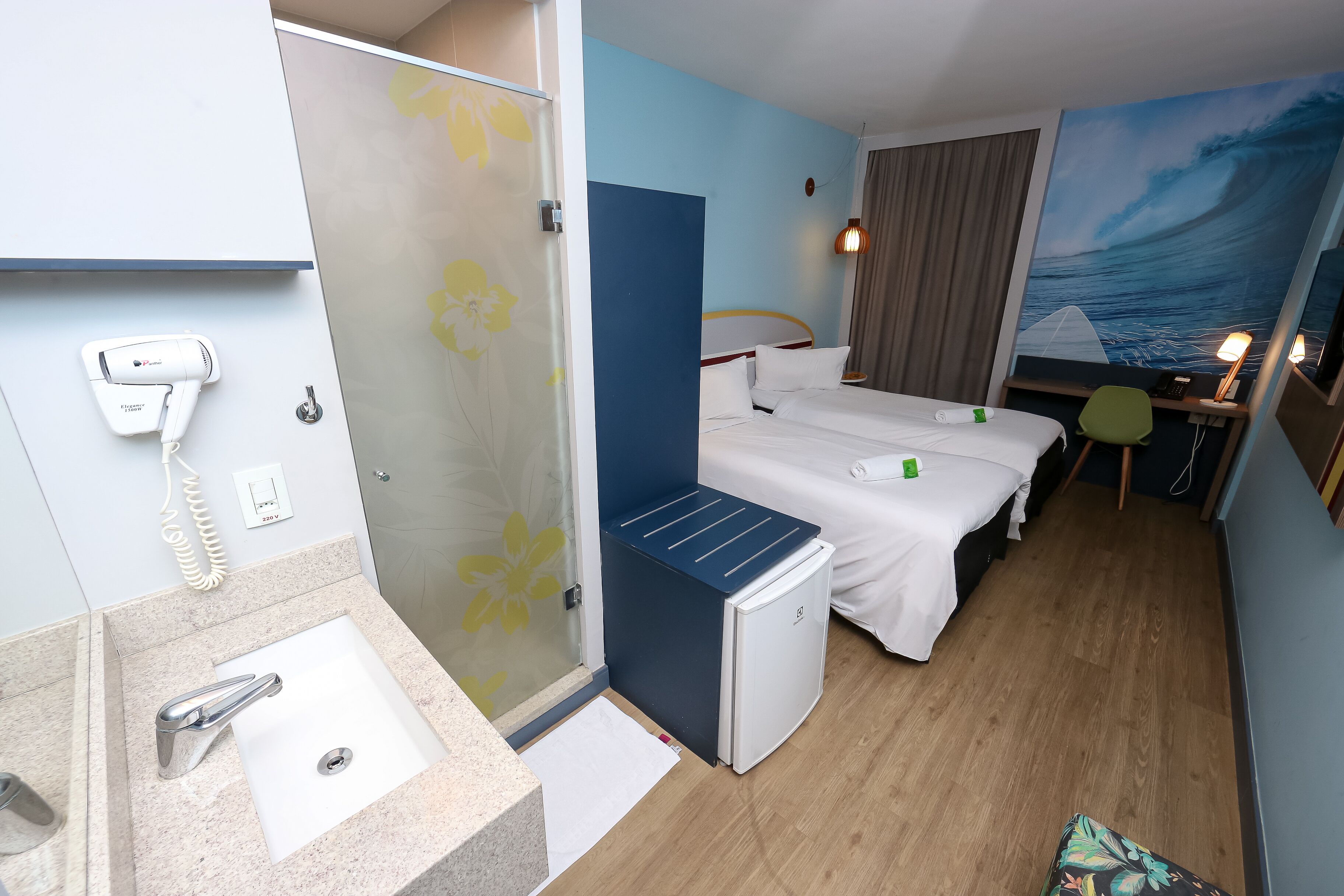 A hotel room with a shower, a sink, a bed, a desk, and a painting of a wave.