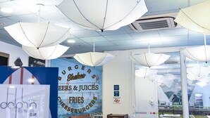A restaurant interior with white tables, chairs, and hanging umbrellas. A menu board is visible with items like burgers, shakes, fries, and cheeseburgers. - Golden Tulip Guarujá (Former Delphin Surf Hotel) (Guarujá)