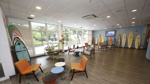 A spacious room with a large window, several chairs, a small table, and surfboards against the wall. - Golden Tulip Guarujá (Former Delphin Surf Hotel) (Guarujá)