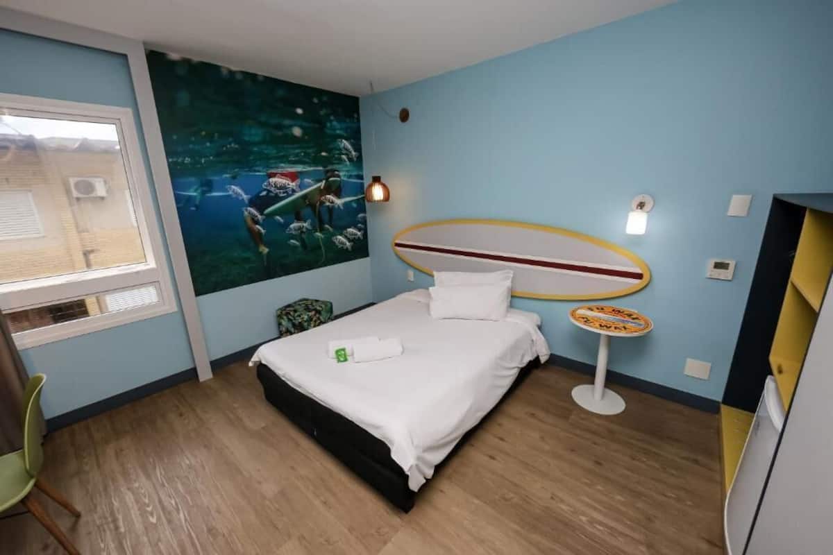 A hotel room with a bed, a wall mural, a small table, and a window.