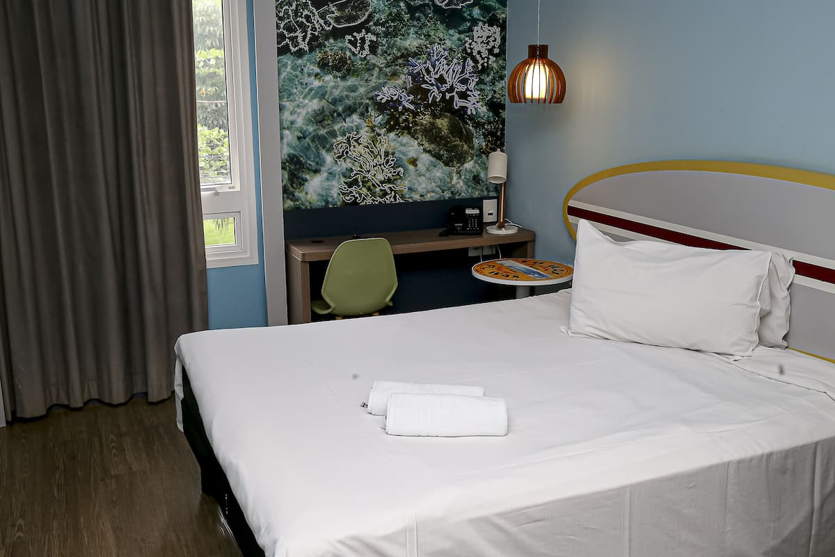 A hotel room with a bed, a desk, a chair, a lamp, and a window with curtains.