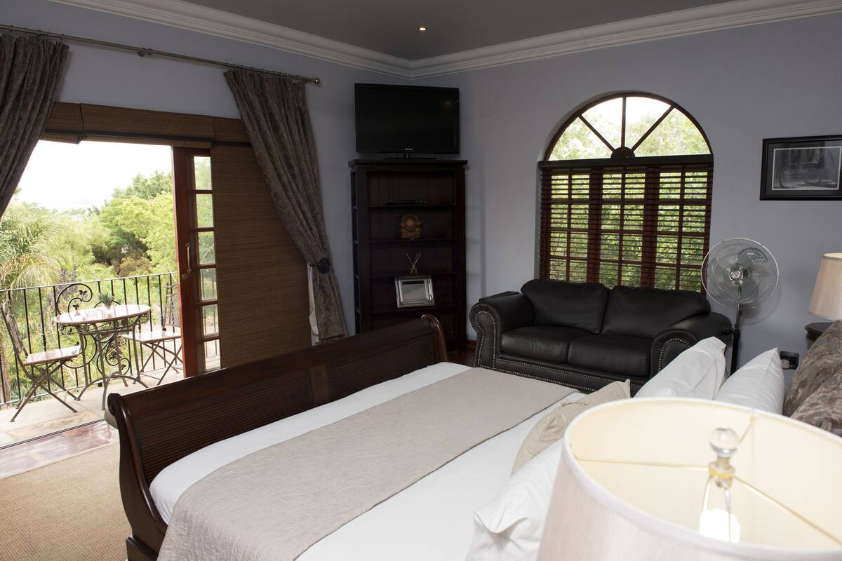 A bedroom with a bed, a television, a sofa, a fan, and a window with a view of greenery.