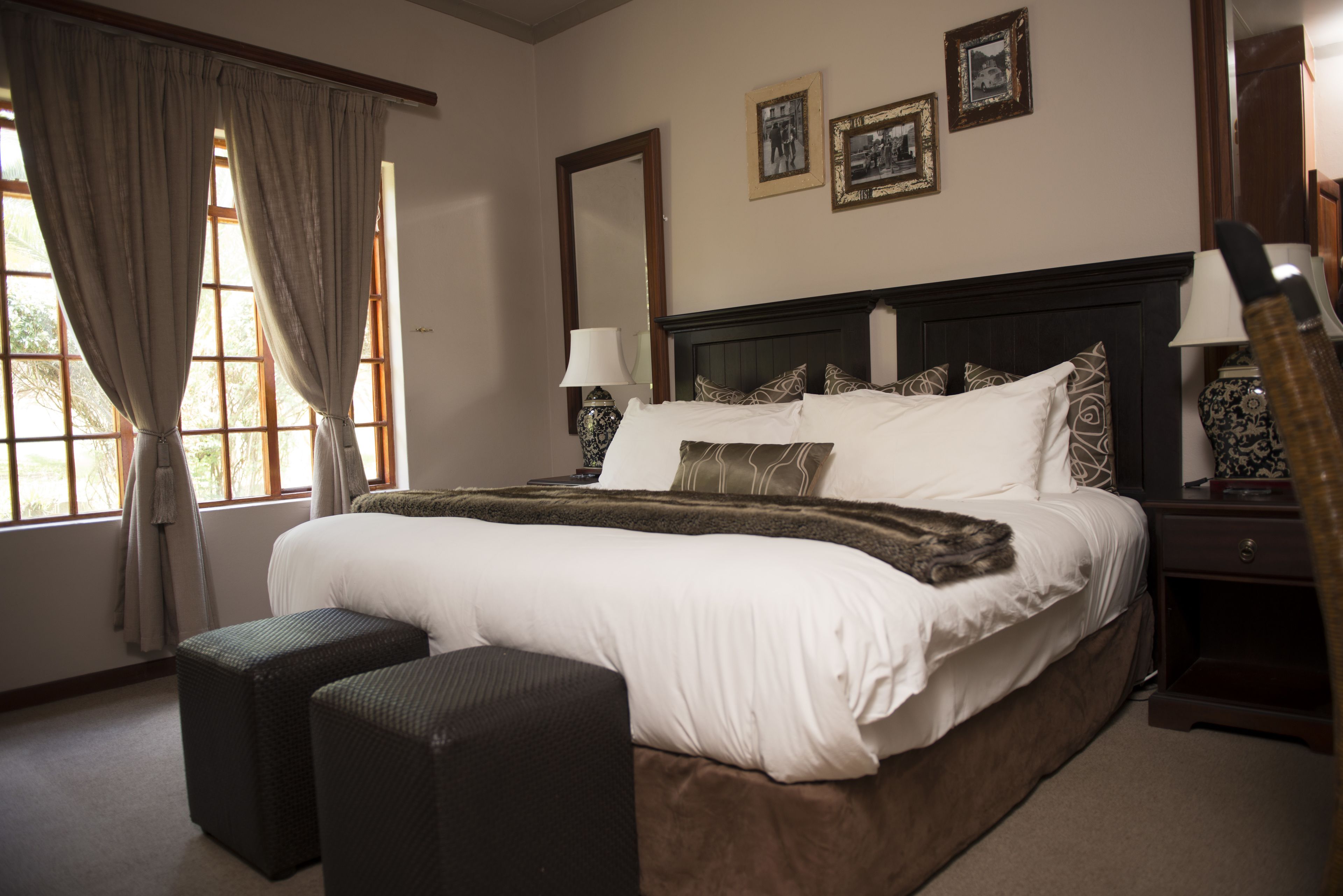 A neatly made bed with white linens and decorative pillows, a bedside table with a lamp, and a large window with curtains.